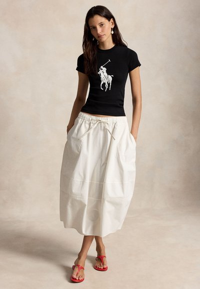 Γυναίκα που φορά μαύρη εφαρμοστή μπλούζα με λευκό σχέδιο παίκτη πόλο, λευκή μίντι φούστα με κορδόνι στη μέση και κόκκινα σαγιονάρα, στέκεται με τα χέρια στις τσέπες.