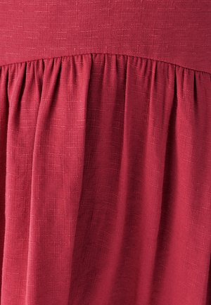 Sarkans audums ar teksturētu austu, kurā ir savāktas detaļas, radot plūstošas krokas. Materiāls izskatās viegls un mīksts.