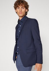 Navy blazer with a tailored fit, single-button closure, and notch lapels. Worn over a dark blue shirt featuring a small white pattern.