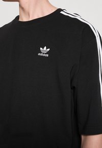 Svart bomullströja med rundhals, som har en vit Adidas-logotyp och två vita ränder längs axlarna. Kort ärm och avslappnad passform.