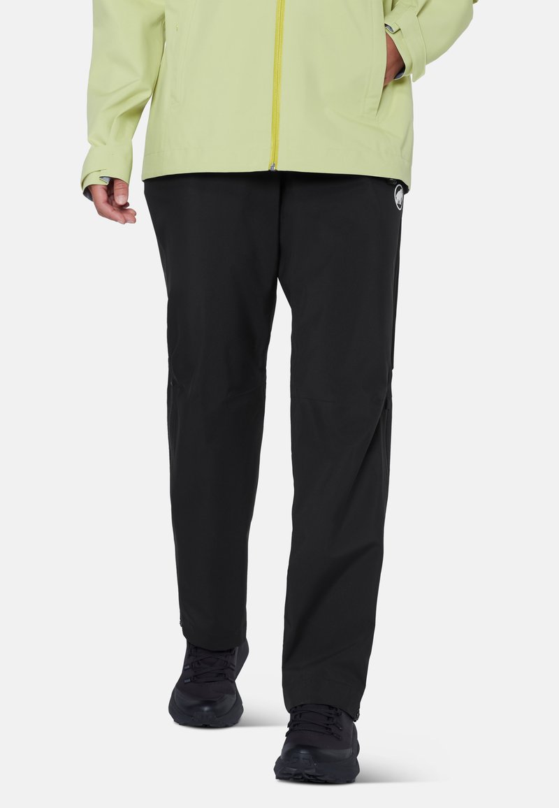 Personne portant un pantalon noir, des chaussures de sport noires et une veste vert clair, les mains dans les poches, marchant vers l'avant sur un fond uni.