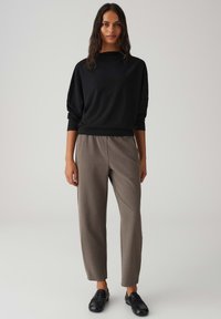 Schwarzer, strukturierter Pullover mit hohem Kragen, kombiniert mit hellbraunen, locker sitzenden Hosen und schwarzen Schnürschuhen.