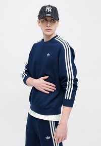 Μπλε σκούρο φούτερ με λευκό σχέδιο τριών ρίγων και μικρό κεντημένο λογότυπο. Συνδυάζεται με παντελόνι σε ίδιο μπλε χρώμα, φορεμένο με λευκό T-shirt από μέσα.