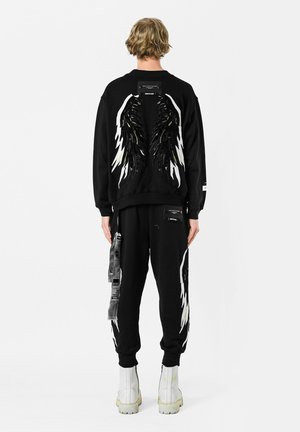 Sudadera negra con diseño de alas que presenta lentejuelas y acentos en blanco contrastantes, combinada con pantalones de chándal negros a juego y botas blancas hasta el tobillo.