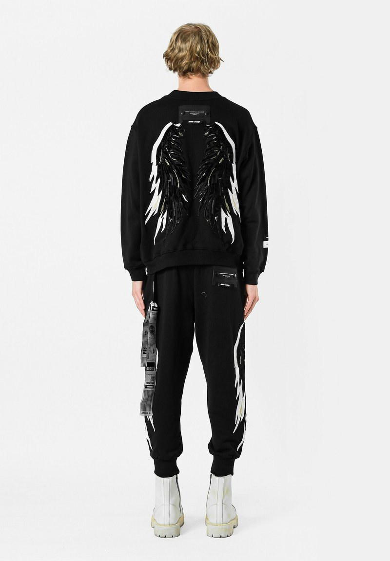 Sudadera negra con diseño de alas que presenta lentejuelas y acentos en blanco contrastantes, combinada con pantalones de chándal negros a juego y botas blancas hasta el tobillo.