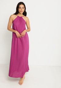 Larga vestido halter en magenta vibrante, con un escote fruncido y tejido fluido. Presenta una silueta suelta y pies descalzos visibles.