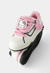 Bílé tenisky s růžovými chlupatými tkaničkami a nášivkou Hello Kitty, s texturovaným svrškem, černými detaily a silnou gumovou podrážkou.