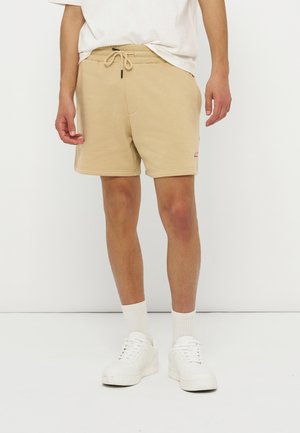 Homme portant un short beige à cordon, des chaussettes blanches montantes, des baskets blanches et un t-shirt blanc, debout devant un fond blanc uni.