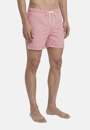 Mann, der rosa-weiß vertikal gestreifte Badeshorts bis zur Mitte des Oberschenkels mit weißem Kordelzug trägt, steht barfuß vor einfarbigem Hintergrund.