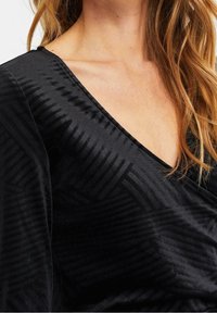 Musta samettinen paita, jossa syvä V-kaula-aukko, teksturoidut vinoraitakuviot ja pehmeä, laskeutuva kangas.