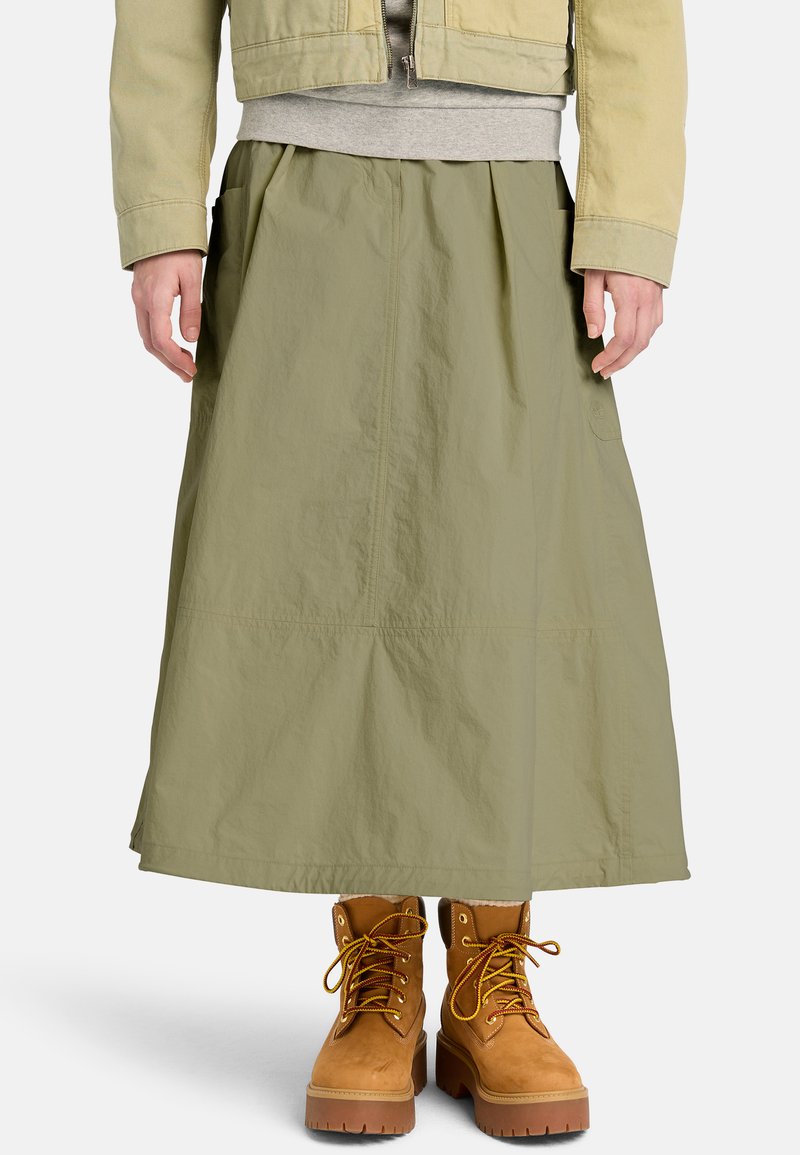 Personne portant une longue jupe vert olive, des bottes lacées couleur tan, un sweat-shirt gris et une veste beige courte sur un fond uni.