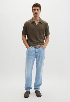 Mężczyzna stoi z rękami w kieszeniach, ubrany w ciemnooliwkową koszulkę polo z krótkim rękawem, jasnoniebieskie dżinsy, beżowy pasek i ciemnobrązowe buty.