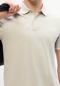 Polo beige en tissu doux avec un col classique, une patte de boutonnage à deux boutons et un petit patch de logo sur la poitrine.