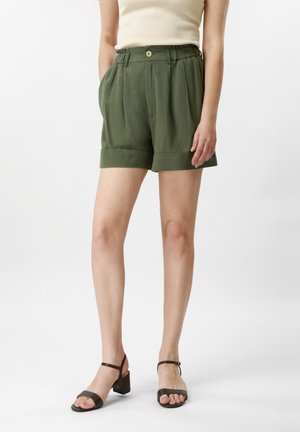 Vrouw die olijfgroene hooggetailleerde geplooide shorts met omgeslagen zomen draagt en een beige mouwloze top, gecombineerd met donkerbruine sandalen met blokhak.