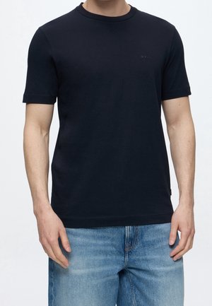 Mężczyzna ubrany w zwykły czarny T-shirt z krótkim rękawem i jasnoniebieskie dżinsy na jednolitym tle.
