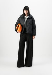 Donna che indossa un giubbotto a vento nero, una camicia bianca, pantaloni neri a gamba larga, mentre tiene una borsa a tracolla arancione, in piedi davanti a uno sfondo bianco.