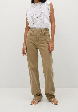 Kobieta ubrana w bezrękawnikowy biały top z koronką ażurową i falbanami, w połączeniu z luźnymi, beżowymi spodniami z wysokim stanem oraz białymi sandałami na ramiączkach.