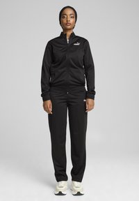 Melnas sporta uzvalks no gludas auduma ar rāvējslēdzēja jaku un elastīgām aprocēm, ar mazu balto Puma logotipu. Saskaņots ar baltām un zaļām sporta kurpēm.