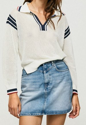 Femme portant un pull blanc perforé avec des rayures bleu marine et une jupe en jean bleu clair, debout les mains détendues le long du corps.