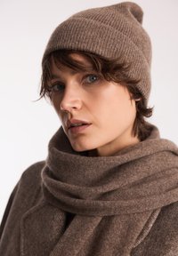 Brązowa dziana czapka z podwiniętym brzegiem, w połączeniu z pasującym brązowym szalikiem i płaszczem. Materiał wygląda na miękki i ma subtelną teksturę.