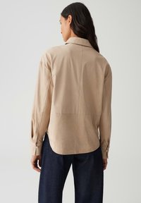 Vrouw in een beige langemouwen shirt en donkerblauwe spijkerbroek, van achteren gezien tegen een effen lichtgrijze achtergrond.