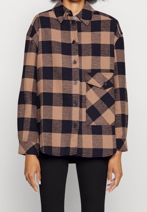 Chemise à carreaux marron et noir avec boutons, manches longues et grande poche avant, portée sur un pantalon noir.
