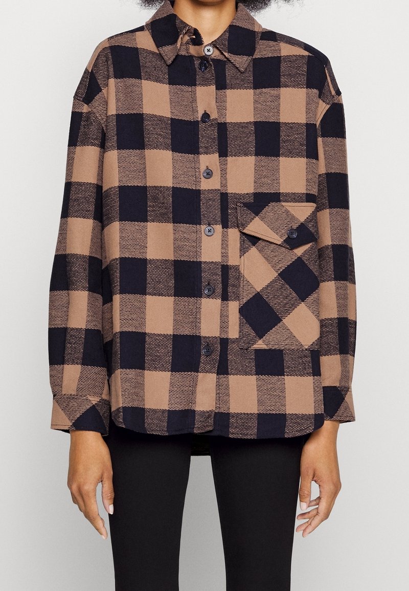 Chemise à carreaux marron et noir avec boutons, manches longues et grande poche avant, portée sur un pantalon noir.