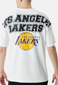 Camiseta blanca con el texto en negrita "LOS ANGELES LAKERS" en negro y un gráfico de baloncesto morado en la parte posterior. Mangas cortas, ajuste relajado.