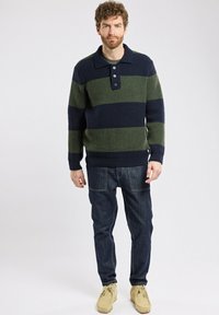 Homme aux cheveux bouclés portant un pull en tricot rayé bleu marine et olive, un jean sombre et des chaussures en daim beige, se tenant contre un fond clair et uni.
