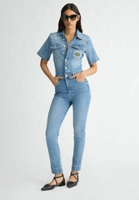Hellblauer Denim-Jumpsuit mit kurzen Ärmeln, tailliertem Schnitt, Knopfverschluss und vorderen Taschen. Getragen mit schwarzen Ballerinas und Sonnenbrille.