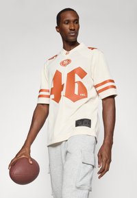 Krémový fotbalový dres s oranžovými akcenty, číslo 46 a logo. Nosit s šedými cargo šortkami. Model drží hnědý fotbal.