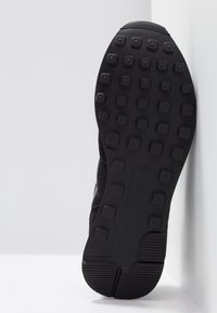 Black shoe sole with square textured tread pattern and horizontal grooves at heel, photographed against white corner background.
