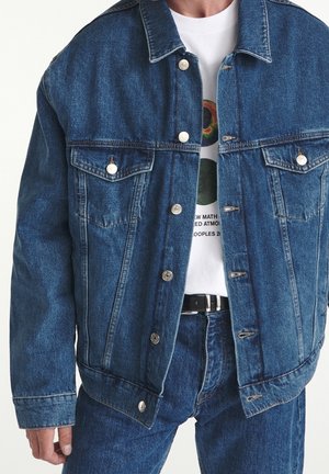 Homme portant une veste en jean bleue déboutonnée et un jean assorti sur un t-shirt blanc avec un motif graphique, ceinture noire visible.