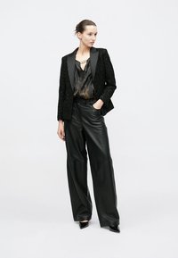 Črni blazer z strukturo in satenastimi reverji, kombiniran s črno bluzo z vzorcem in širokimi hlačami iz usnja. Celoten videz dopolnjujejo sijoči črni čevlji s peto.