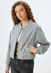 Graue, taillierte Bomberjacke mit Druckknopfverschluss, geripptem Kragen und zwei Seitentaschen; aus weichem, strukturiertem Stoff gefertigt.