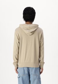 Sweat à capuche beige à manches longues, avec une poche kangourou et une texture douce. Le design présente une coupe décontractée et des poignets côtelés.