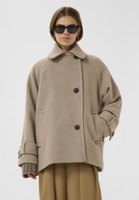 Beige wollen coat met een knoopsluiting aan de voorkant, brede kraag en grote zakken. Heeft oversized mouwen met verstelbare manchetten en een relaxte pasvorm.