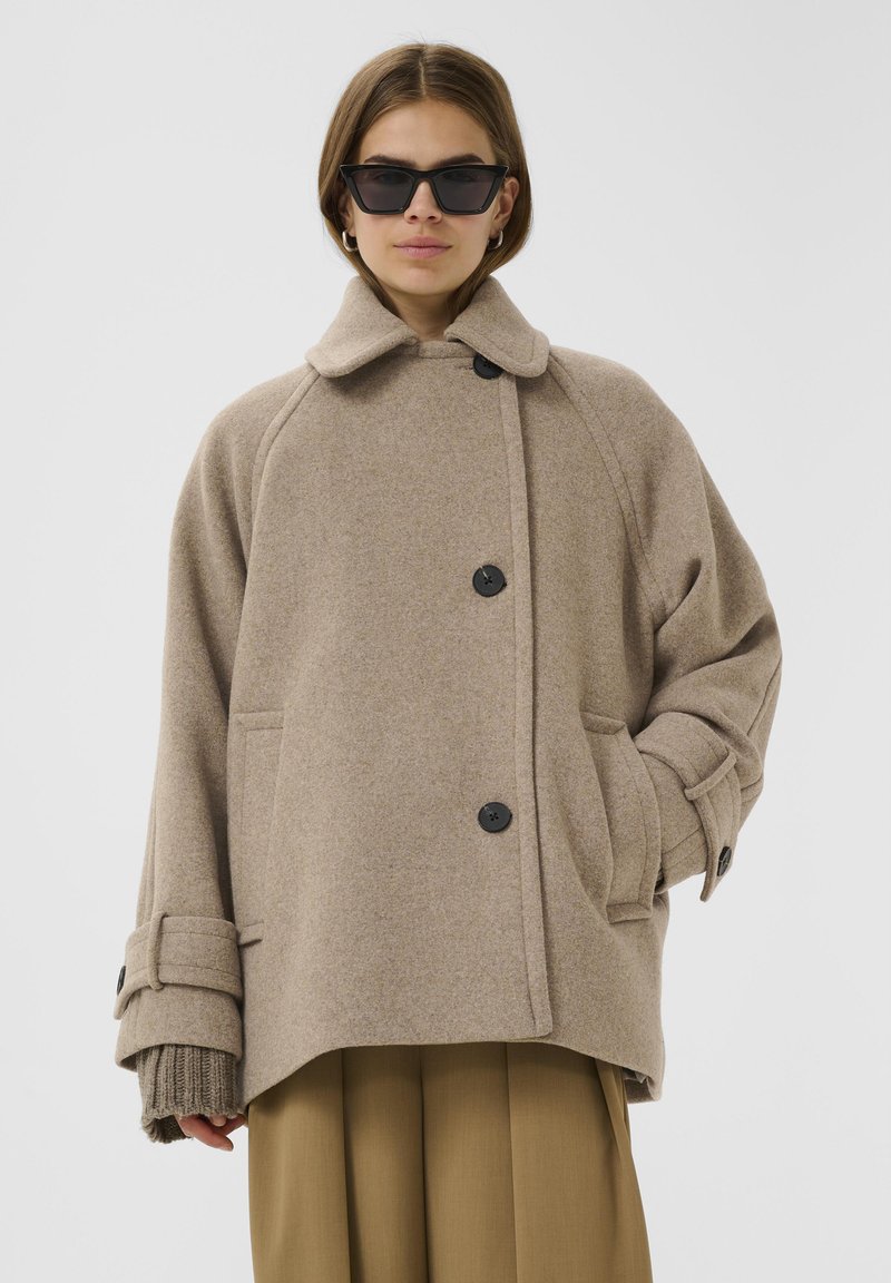 Beige wollen coat met een knoopsluiting aan de voorkant, brede kraag en grote zakken. Heeft oversized mouwen met verstelbare manchetten en een relaxte pasvorm.