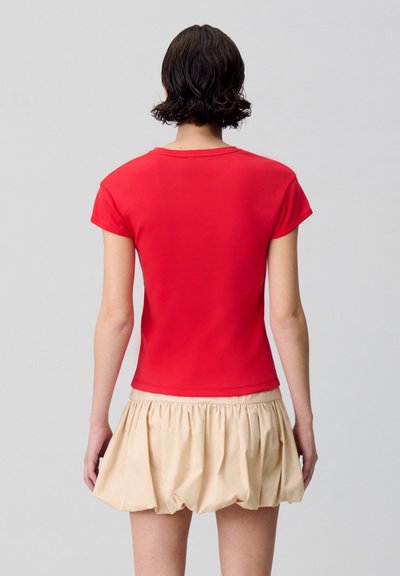 T-shirt rossa in cotone a maniche corte e scollatura rotonda, abbinata a una gonna beige a pieghe, vista da dietro.