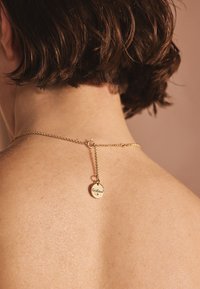 Zelta ķēdes kaklarota ar piekariņu, kurā redzams logotipa elements, atrodas uz kailas ādas. Ķēdei ir smalka saišu konstrukcija un aizdare.