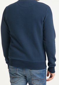 Granatowa bluza z dekoltem w kształcie zaokrąglonego kołnierza, wykonana z miękkiego materiału, z długimi rękawami i ściągaczami na mankietach, zestawiona z jasnoniebieskimi dżinsami.