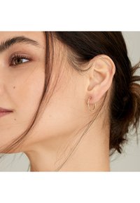 Guld hoops ørering med et tyndt, struktureret design. Lille accent nær lukningen tilføjer detalje. Båret i venstre øre, komplementeret af løst hår.