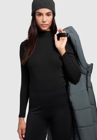 Schwarzes Langarmshirt mit Stehkragen, taillierte Passform, kombiniert mit schwarzen Leggings und grauer Steppweste. Das Modell trägt eine graue Strickmütze.