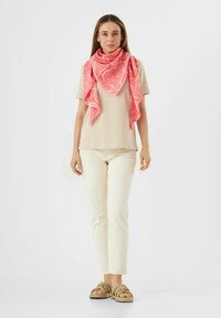 Roze sjaal met bloemenpatroon om de nek, gecombineerd met een beige shirt en crèmekleurige broek, aangevuld met geknoopte sandalen.