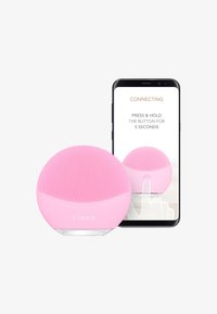 Dispositif de nettoyage facial en silicone rose, avec une surface texturée et une forme arrondie, à côté d'un smartphone affichant un écran de connexion.