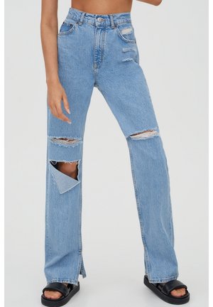 Jean en denim déchiré bleu clair avec taille haute, jambes évasées et plusieurs patches vieillissants. Présente une fermeture à bouton et des détails d'ourlet effilochés.