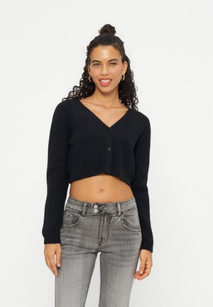 Femme aux cheveux bouclés portant un cardigan noir court et un jean gris taille haute, souriante sur un fond blanc.