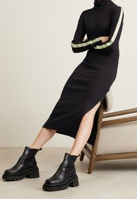 Svart ribbat turtleneck-klänning med långa ärmar, med gröna textaccenter och slits på sidan. Matchad med kraftiga svarta läderst övlar vid ankeln.