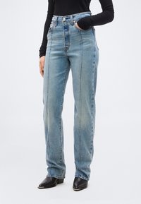 Γυναίκα με ψηλόμεσα μπλε τζιν με εμφανή μπροστινά ραφές, σε συνδυασμό με μαύρα μυτερά μποτάκια και μαύρο μακρυμάνικο τοπ.