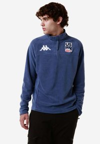 Pullover in fleece blu navy con zip a quarti e logo Kappa e emblema degli Stati Uniti. Presenta una consistenza morbida e maniche lunghe, progettato per il comfort.